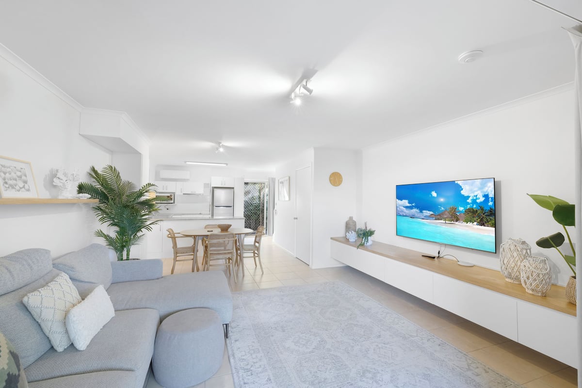A modern living room with a grey sofa, a flat-screen TV, and a dining area with a table and chairs.