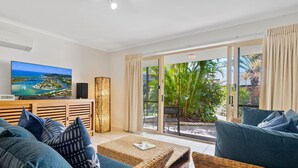 Una sala de estar moderna con un televisor de pantalla plana grande, asientos cómodos y un ventanal corredero que da a un jardín tropical.