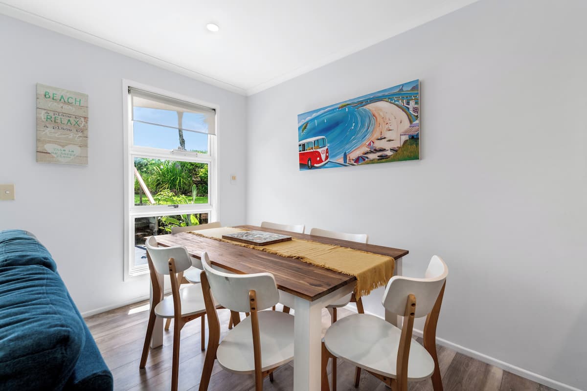 Un comedor con una mesa de madera y sillas blancas, un cuadro de una playa y una ventana con vistas a la vegetación.