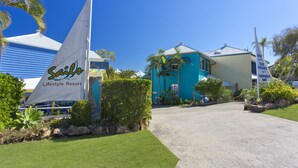 Un bâtiment situé sur la côte, avec un voilier, des palmiers et une enseigne indiquant « Sailo Lifestyle Resort ».