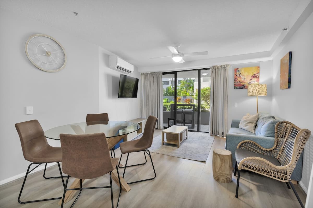 A modern living room with a glass dining table, brown chairs, a blue sofa, a wicker chair, a floor lamp, and a TV mounted on the wall.