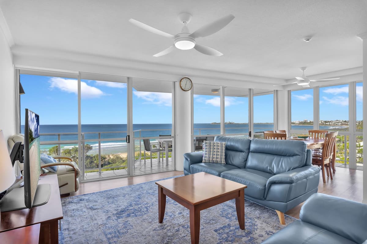 A spacious living room with a large window overlooking the ocean, a ceiling fan, and a wooden coffee table.