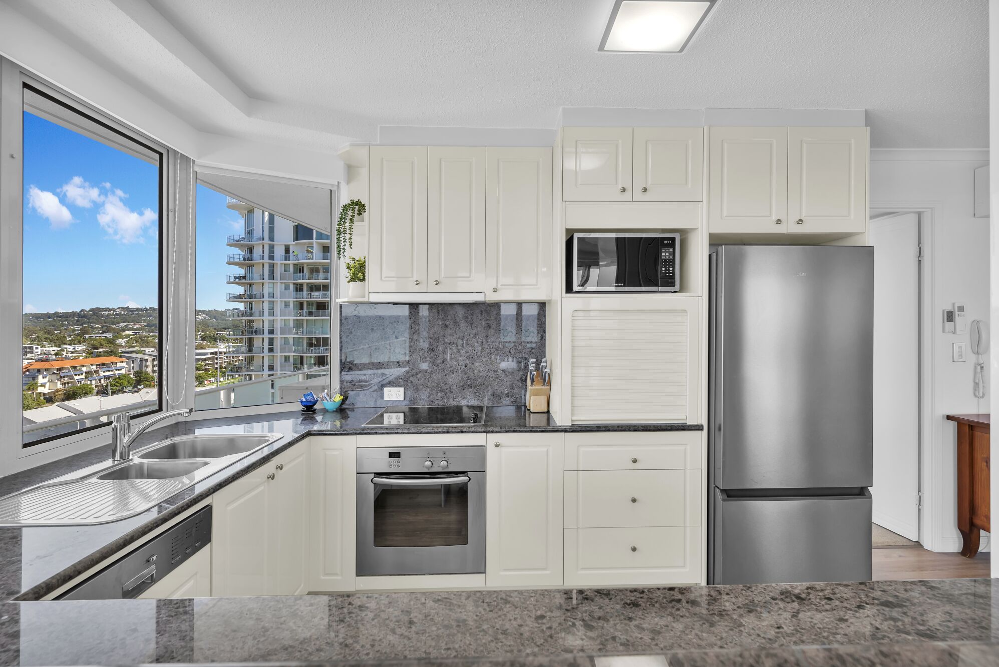 A modern kitchen with white cabinets, stainless steel appliances, a built-in oven, and a large window offering a city view.