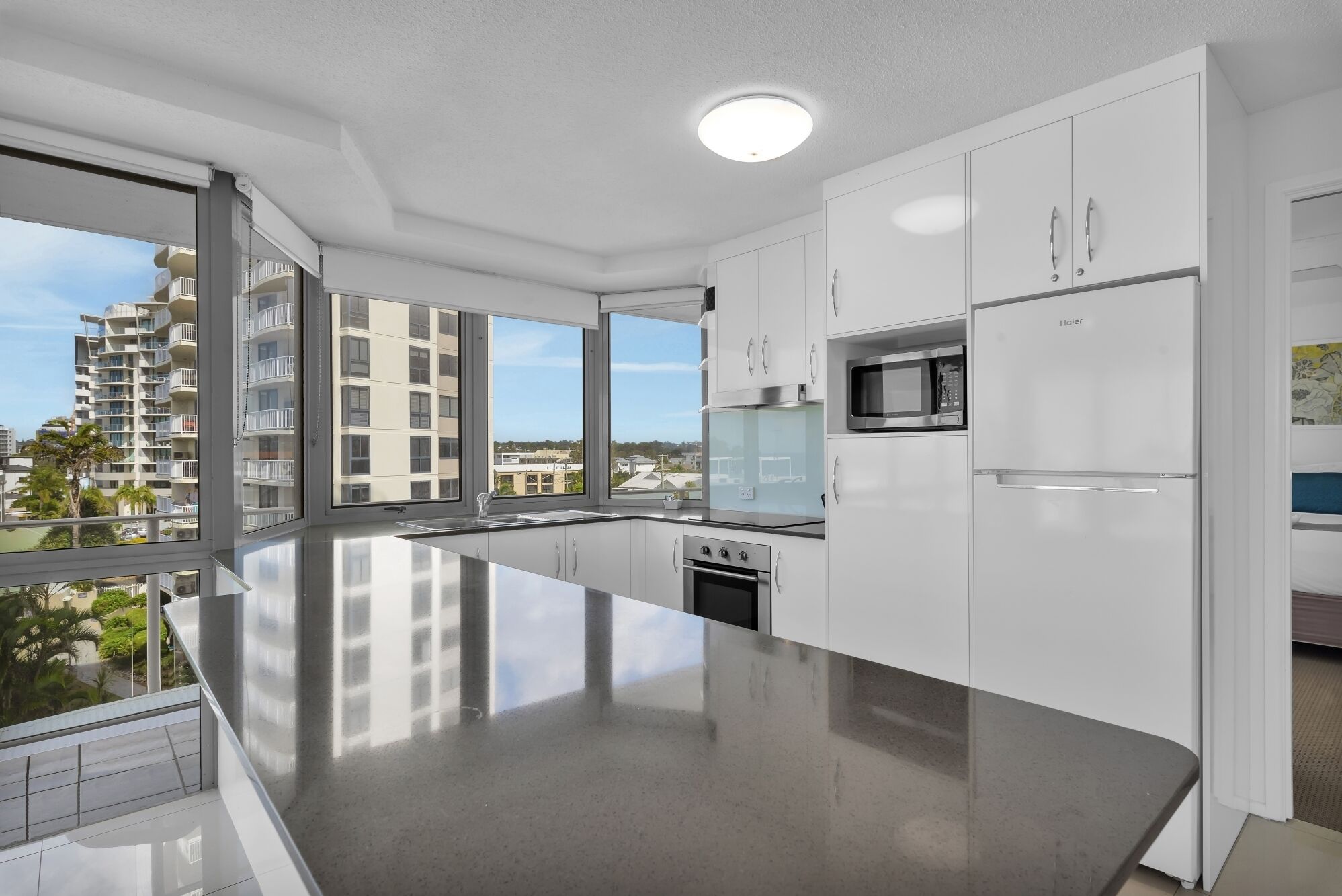 A modern kitchen with white cabinets, a stainless steel refrigerator, and a large window offering a view of buildings and greenery.