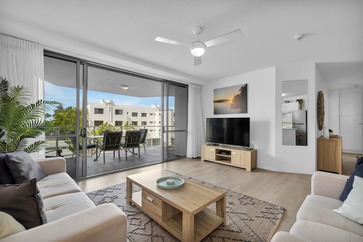 A modern living room with a large TV, wooden coffee table, and a balcony with a view of buildings.