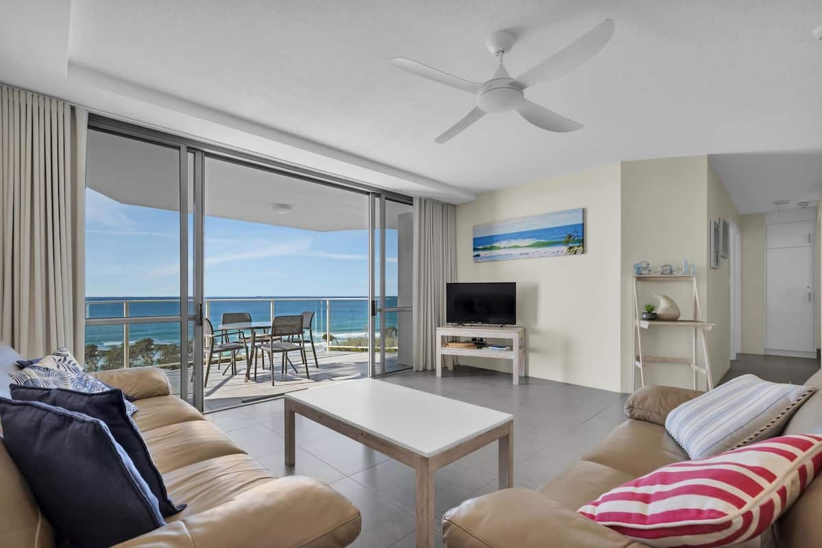 A modern living room with a large sliding glass door leading to a balcony with a view of the ocean. There is a flat-screen TV on a wooden stand, a beige sofa, and a coffee table.