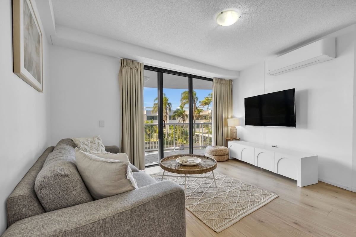 A modern living room with a sofa, a round table with a bowl, a TV mounted on the wall, and a balcony with a view of palm trees.