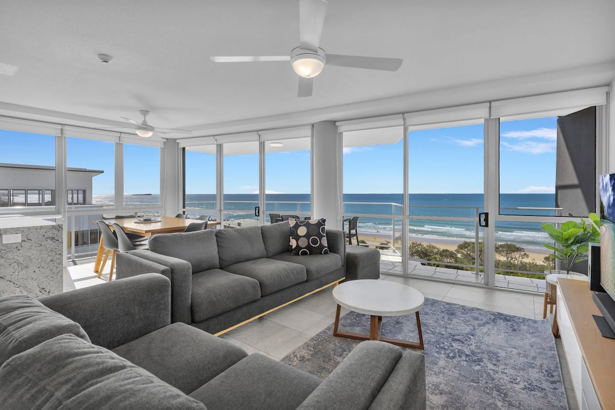 A modern living room with a large gray sofa, a round coffee table, and a TV. The room has large windows offering a view of the ocean.