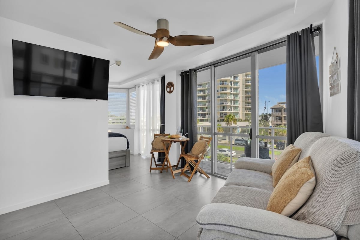A modern living room with a large flat-screen TV, a ceiling fan, and a sofa with cushions. There is a dining area with a table and chairs, and a balcony with a view of the city.