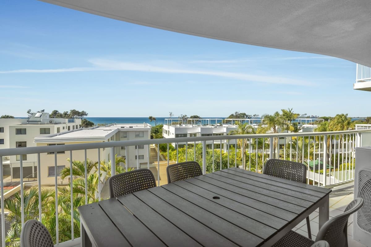 A balcony with a table and chairs, overlooking a coastal area with buildings and palm trees.