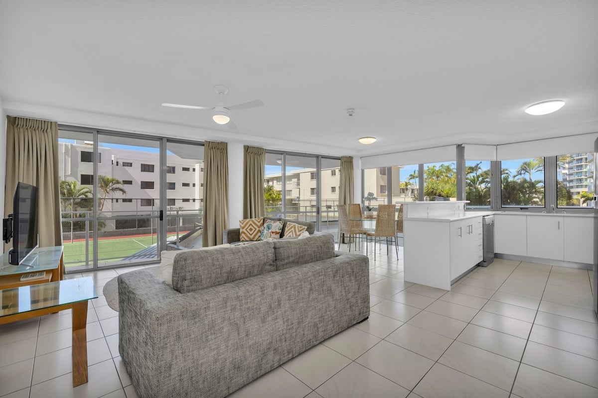 A modern apartment with a living area, dining space, and kitchen. It features large windows, a ceiling fan, and a view of a sports field and buildings.