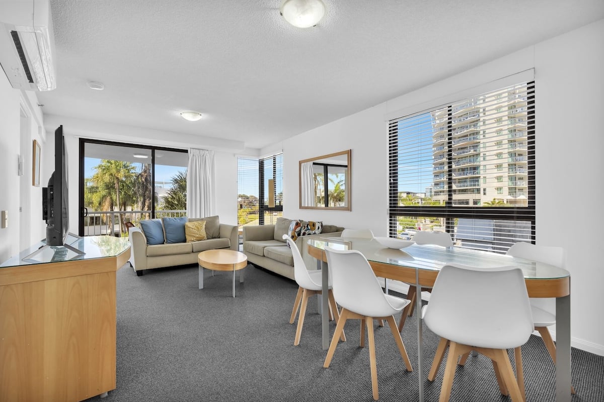 A modern living room with a sofa, a dining table with chairs, a TV on a stand, and large windows with blinds.