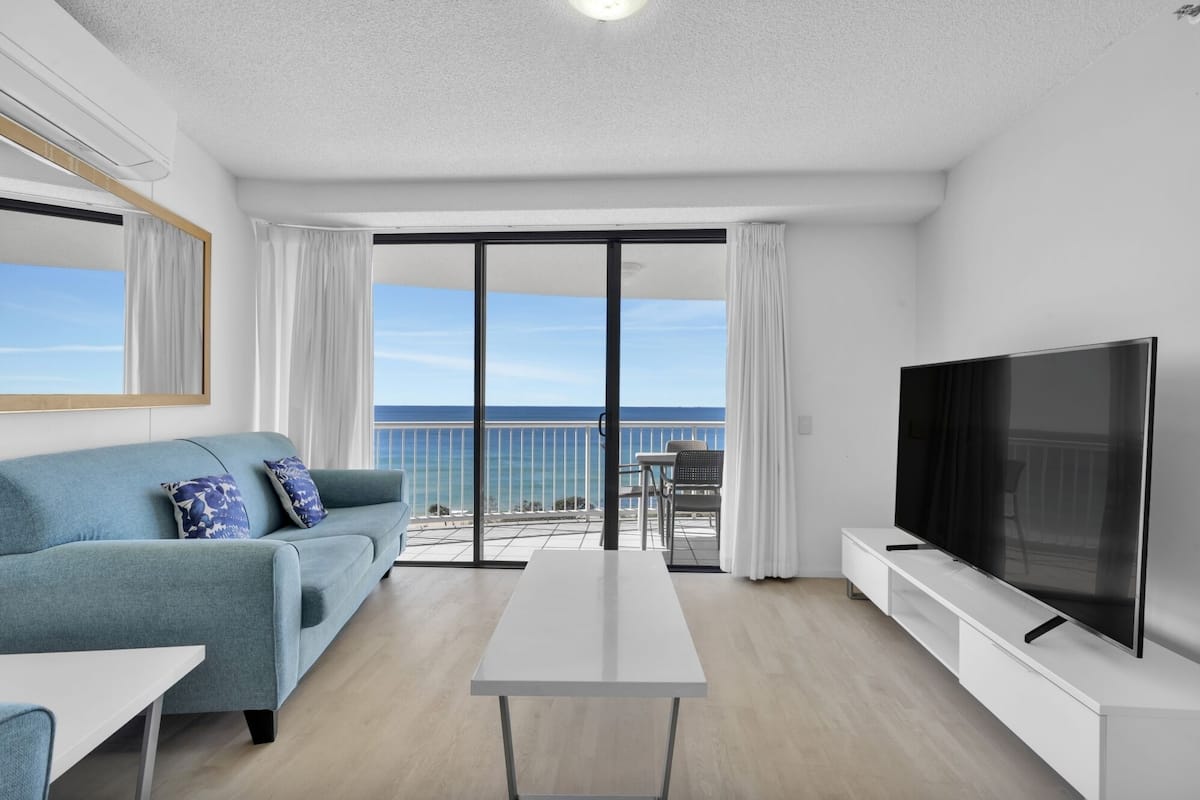 A modern living room with a sofa, a TV, and a balcony with a view of the ocean.