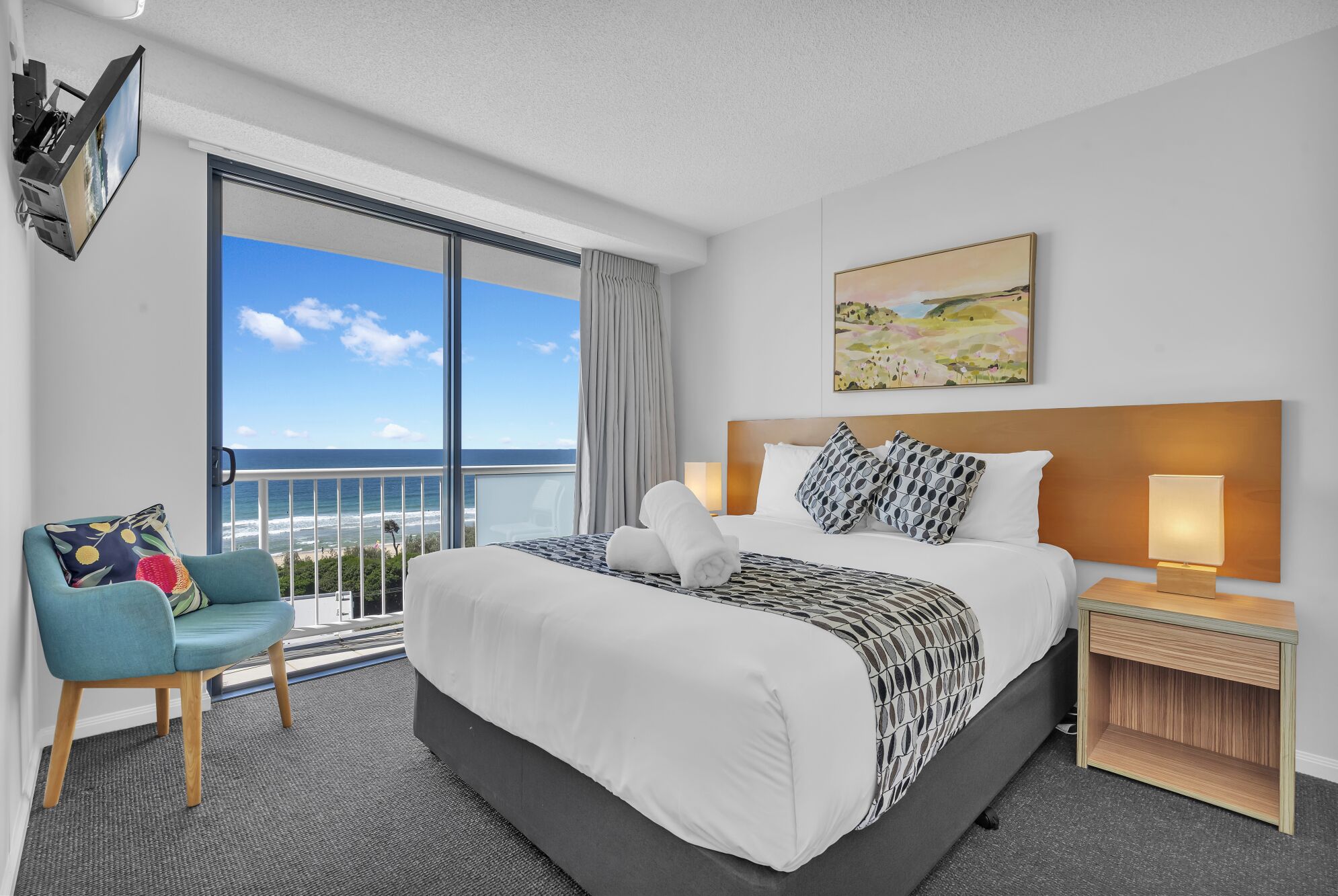 Quarto de hotel com uma cama grande, uma cadeira, uma mesinha de cabeceira com abajur, um quadro na parede e vista para o oceano.
