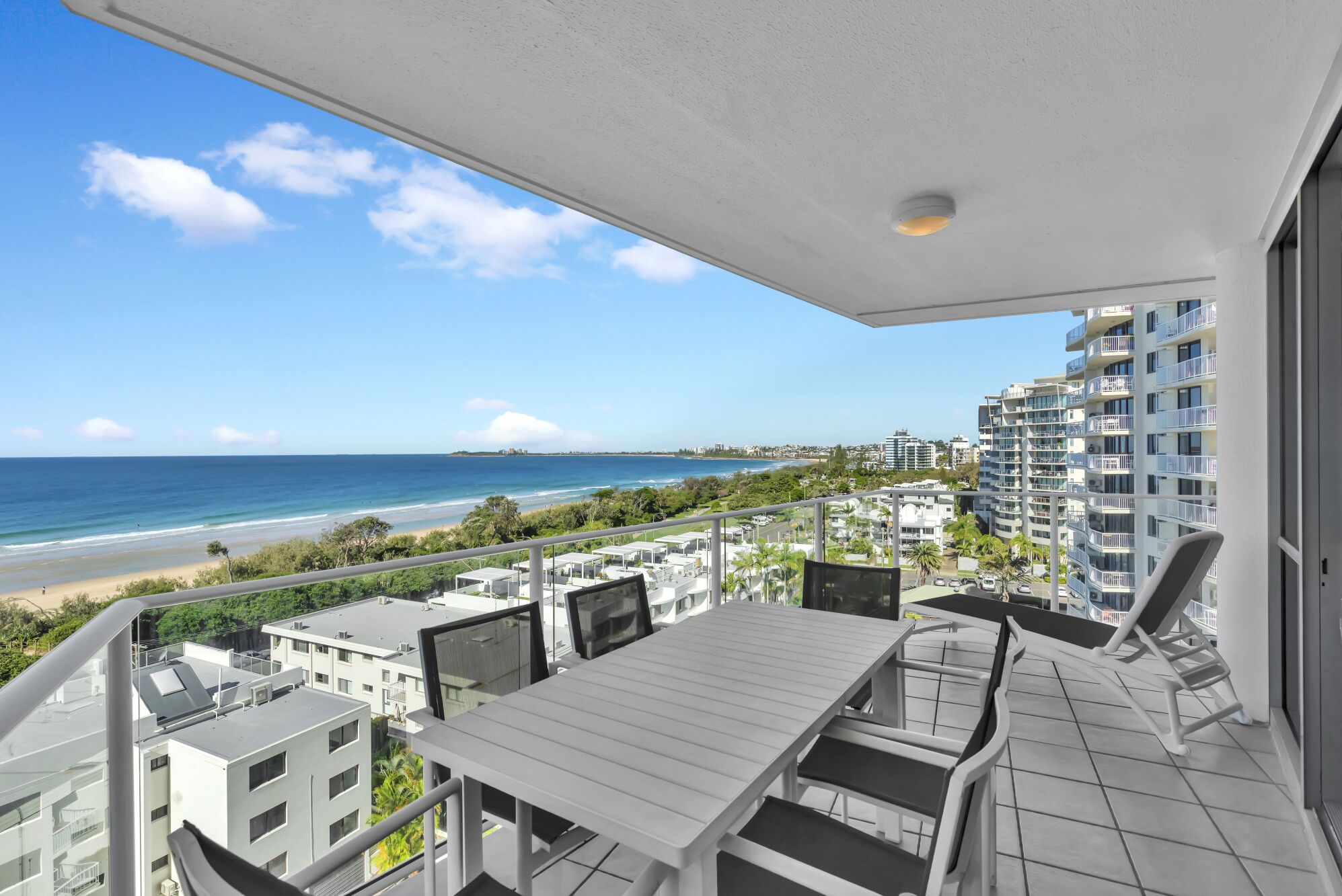 Uma varanda com vista para a praia e oceano, edifícios modernos ao fundo e uma mesa branca com cadeiras.