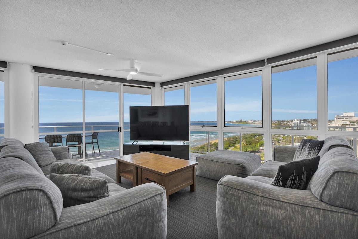 A modern living room with a large TV, grey sofas, a wooden coffee table, and a balcony with a view of the sea.
