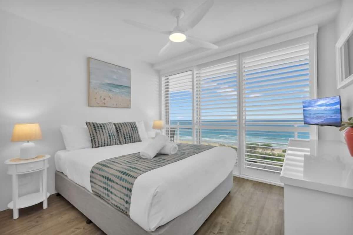 A bedroom with a bed, a ceiling fan, a TV, and a view of the ocean.