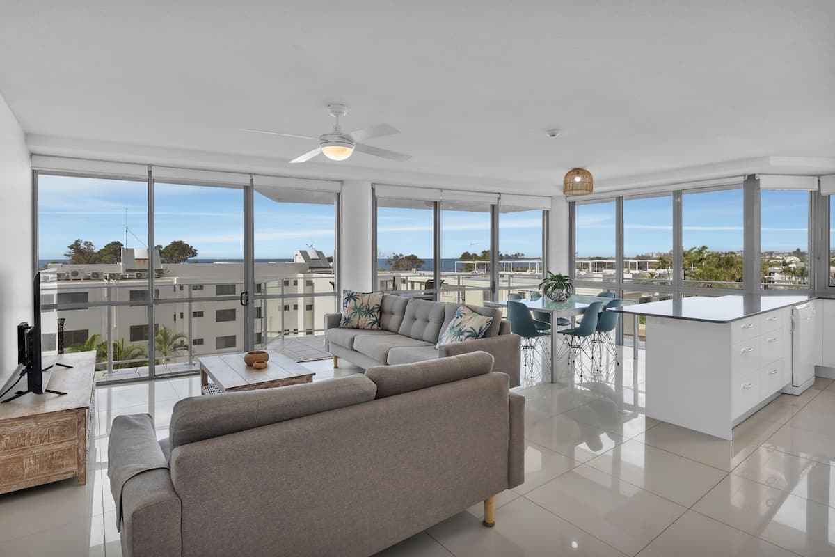 A spacious living area with a sofa, dining table, and chairs, large windows offering a view of the cityscape.