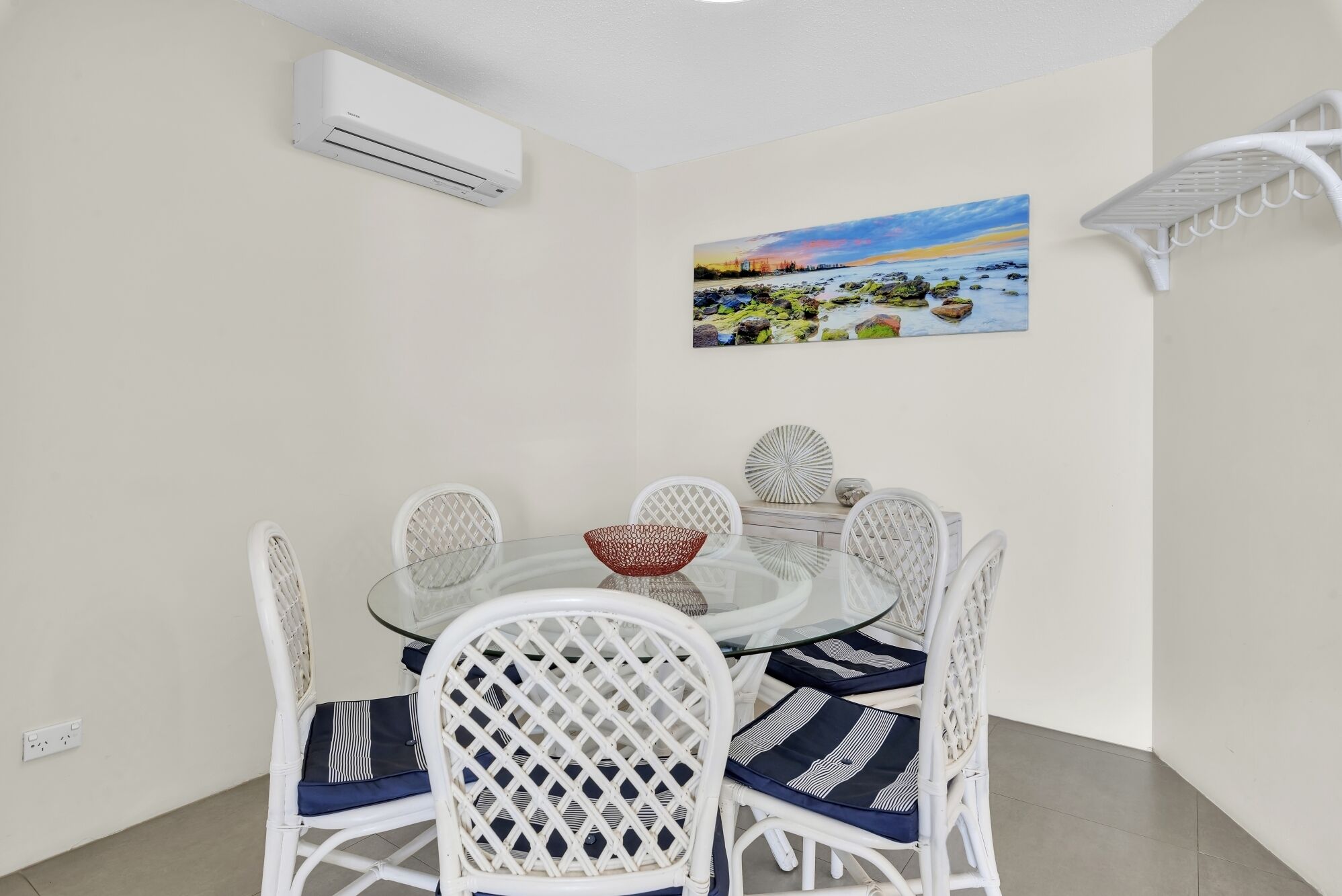 A dining area with a glass table, white wicker chairs, and a painting of a coastal scene.