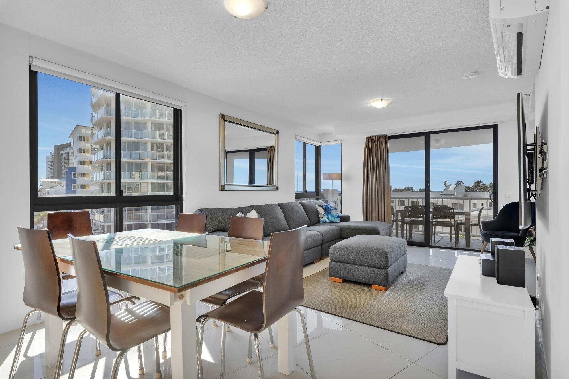 Una sala de estar moderna con una mesa de comedor de vidrio, sofá gris y un balcón con vistas a la ciudad.