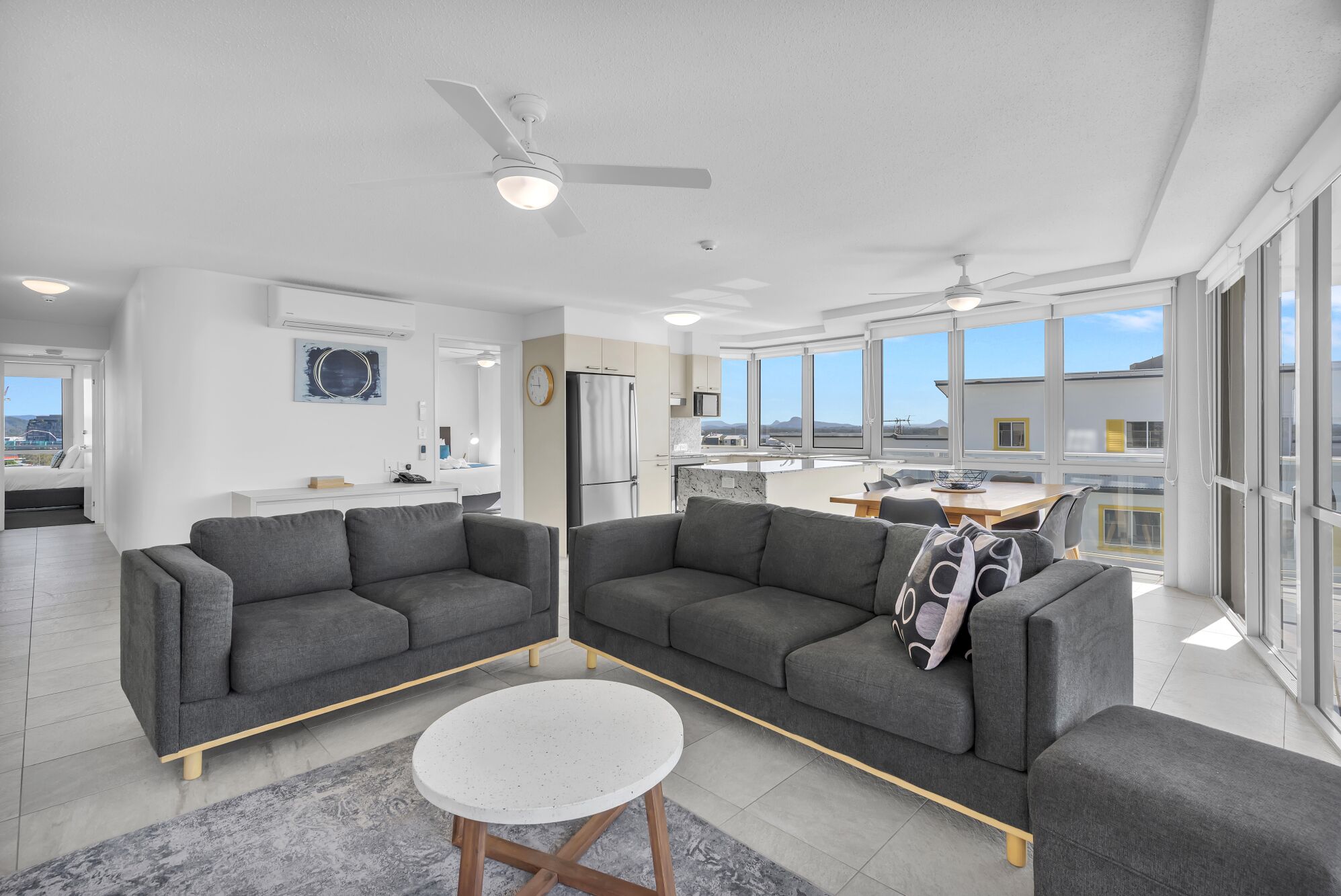 A modern living room with grey sofas, a round coffee table, and a view of the kitchen and outside.