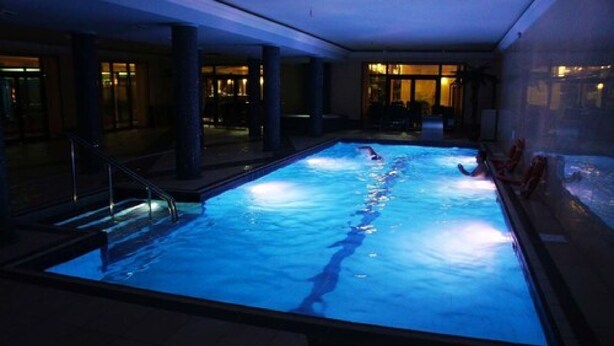 An indoor swimming pool with a clear water path, surrounded by dark-toned walls and columns, and a staircase leading to an upper level. - Dobosz (Police)