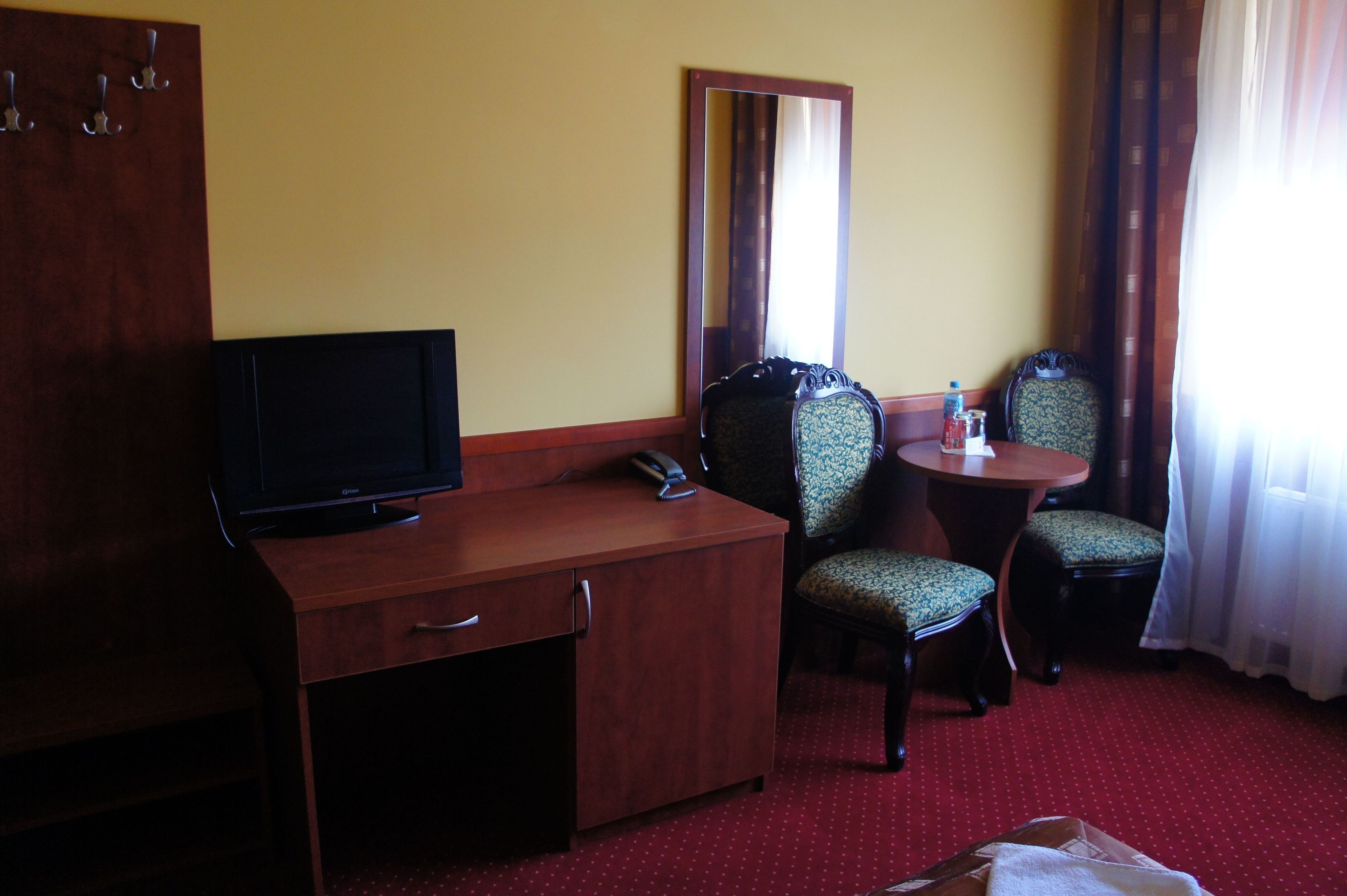 Una camera d'hotel con una scrivania in legno, una televisione, due poltrone a fantasia, un tavolino con una bottiglia e un bicchiere, e uno specchio a parete.
