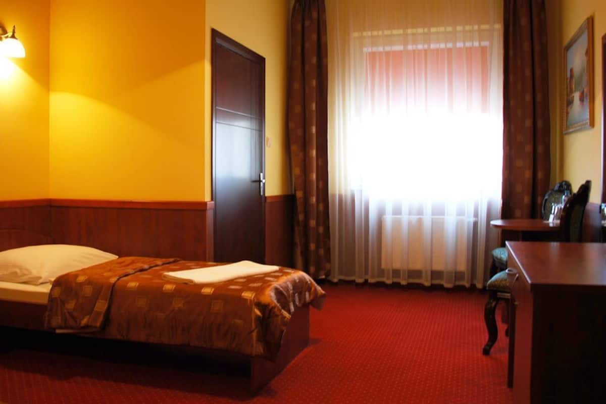 Quarto de hotel com cama, escrivaninha, cadeira, janela com cortinas e luminária na parede.