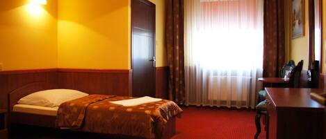 A hotel room with a bed, a desk, a chair, a window with curtains, and a wall-mounted lamp.