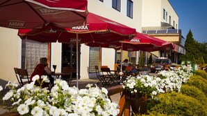Outdoor seating with red umbrellas, white flowers, and a building with multiple windows. - Dobosz (Police)