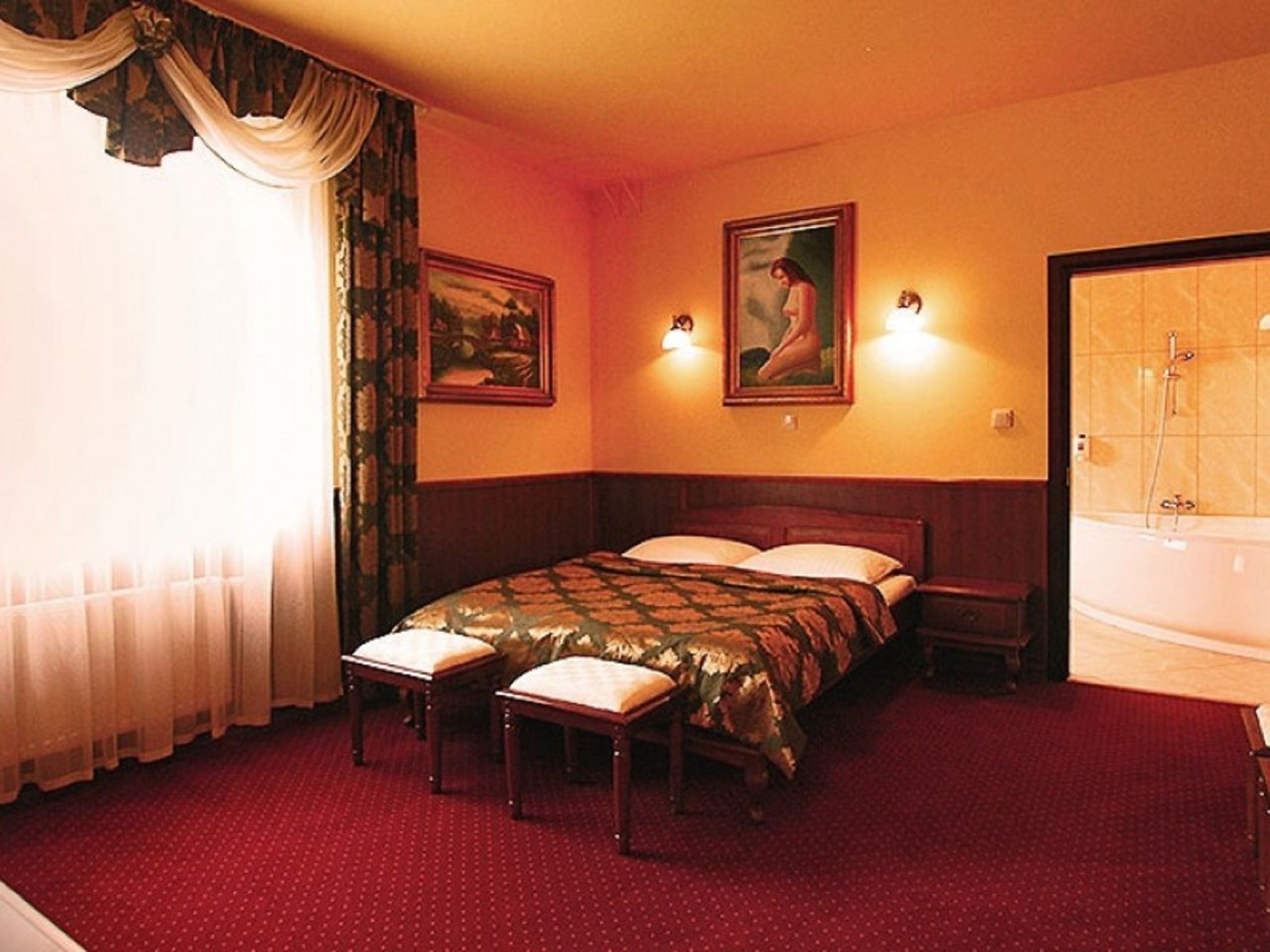 A hotel room with a large bed, two bedside tables, a nightstand, a window with curtains, a painting on the wall, and a bathroom with a bathtub.
