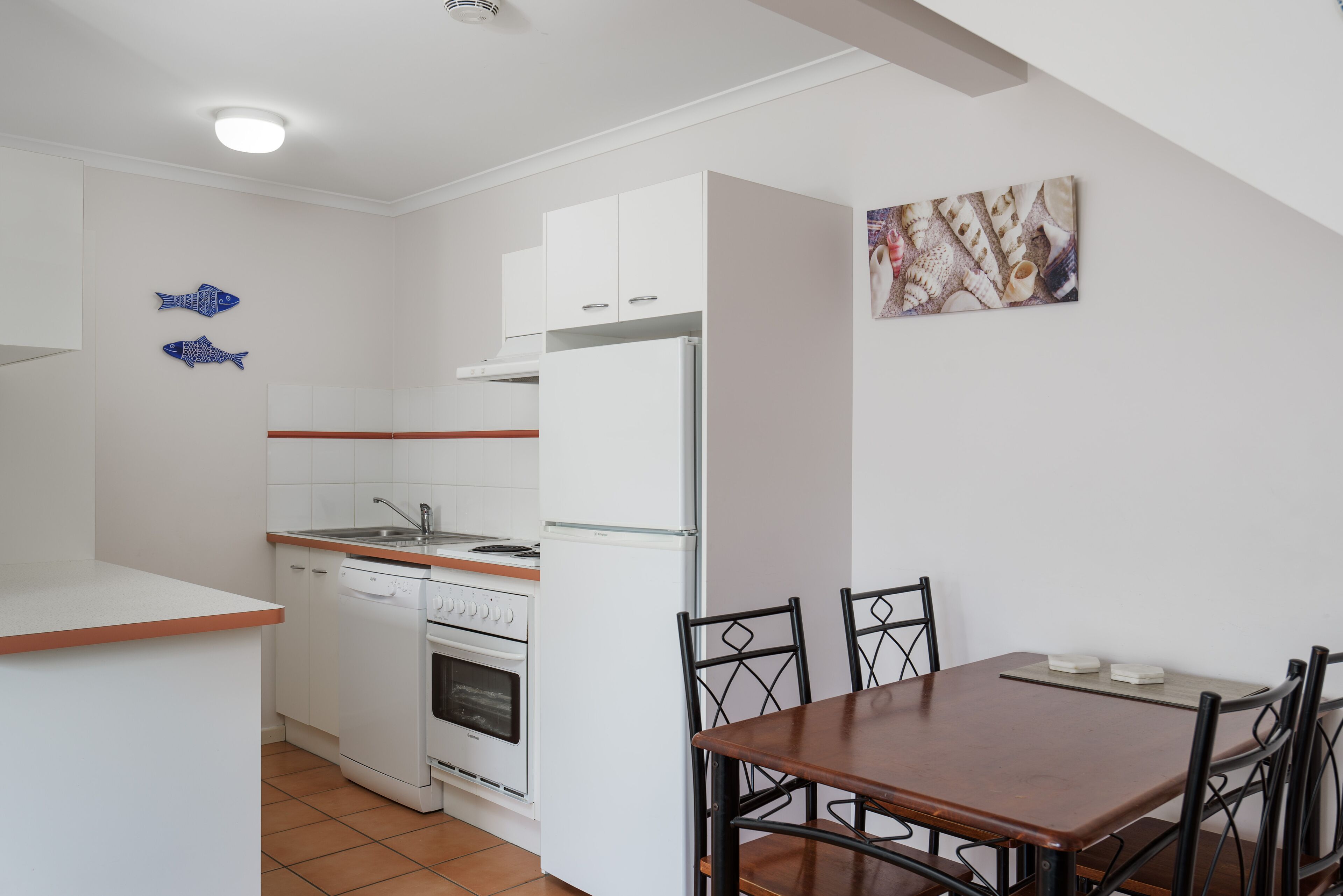 Cocina compacta con armarios blancos, un refrigerador blanco, lavavajillas y estufa. Hay una mesa de comedor con cuatro sillas.