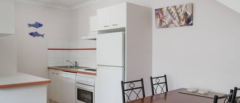 Cocina compacta con armarios blancos, un refrigerador blanco, lavavajillas y estufa. Hay una mesa de comedor con cuatro sillas.