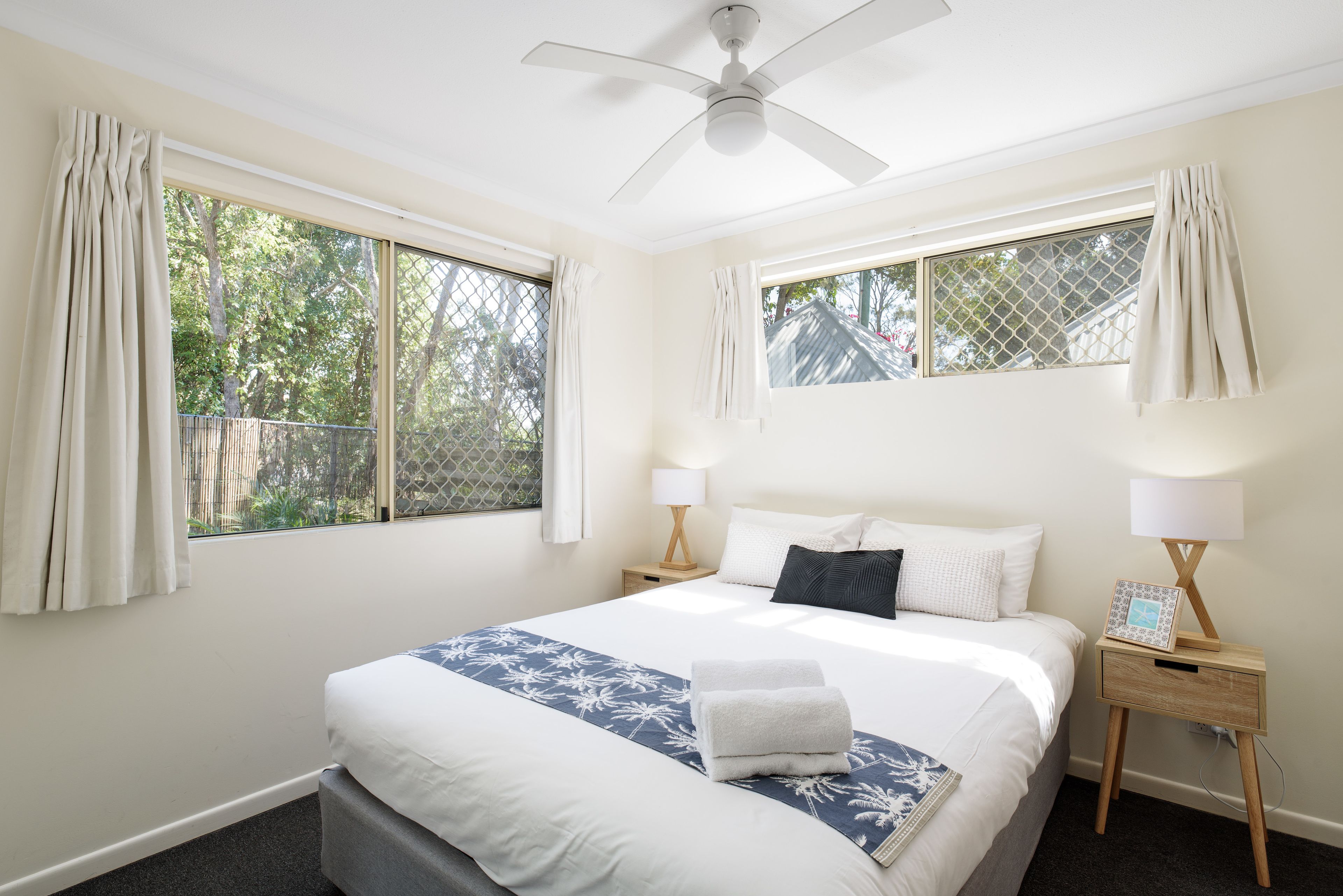 A bedroom with a bed, two bedside tables with lamps, a ceiling fan, and large windows with curtains.