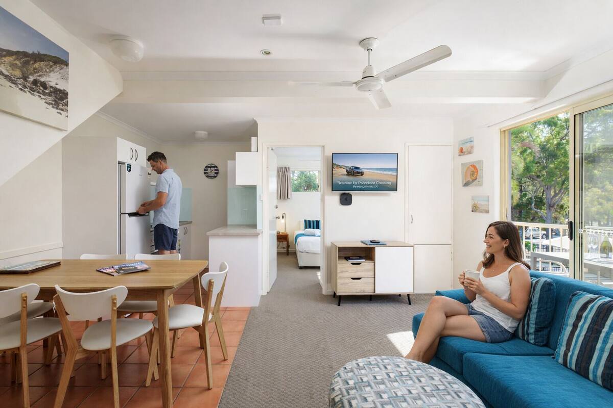 A modern living room with a sofa, a dining area with chairs, a kitchen with a person cooking, and a TV on the wall.