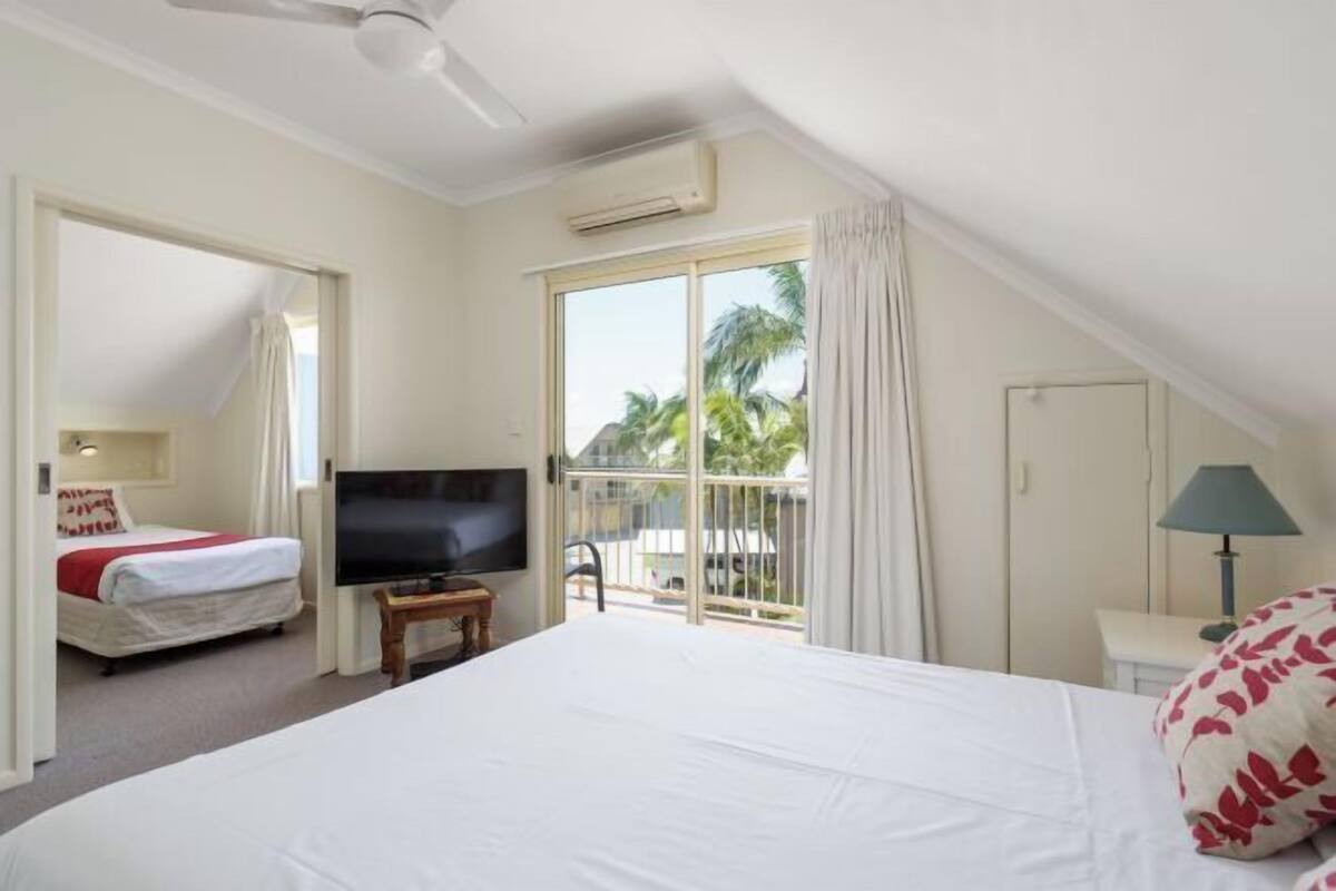 A hotel room with a large bed, a TV, a balcony with a view of palm trees, and an air conditioning unit.