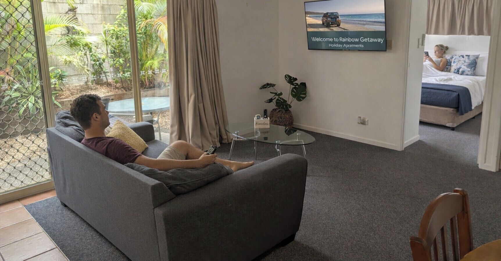 A living room with a sofa, a coffee table, a TV, and a bed in the background.