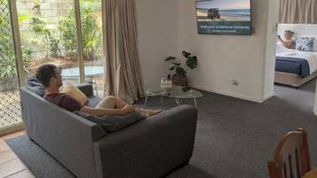 A living room with a sofa, a coffee table, a TV, and a bed in the background.