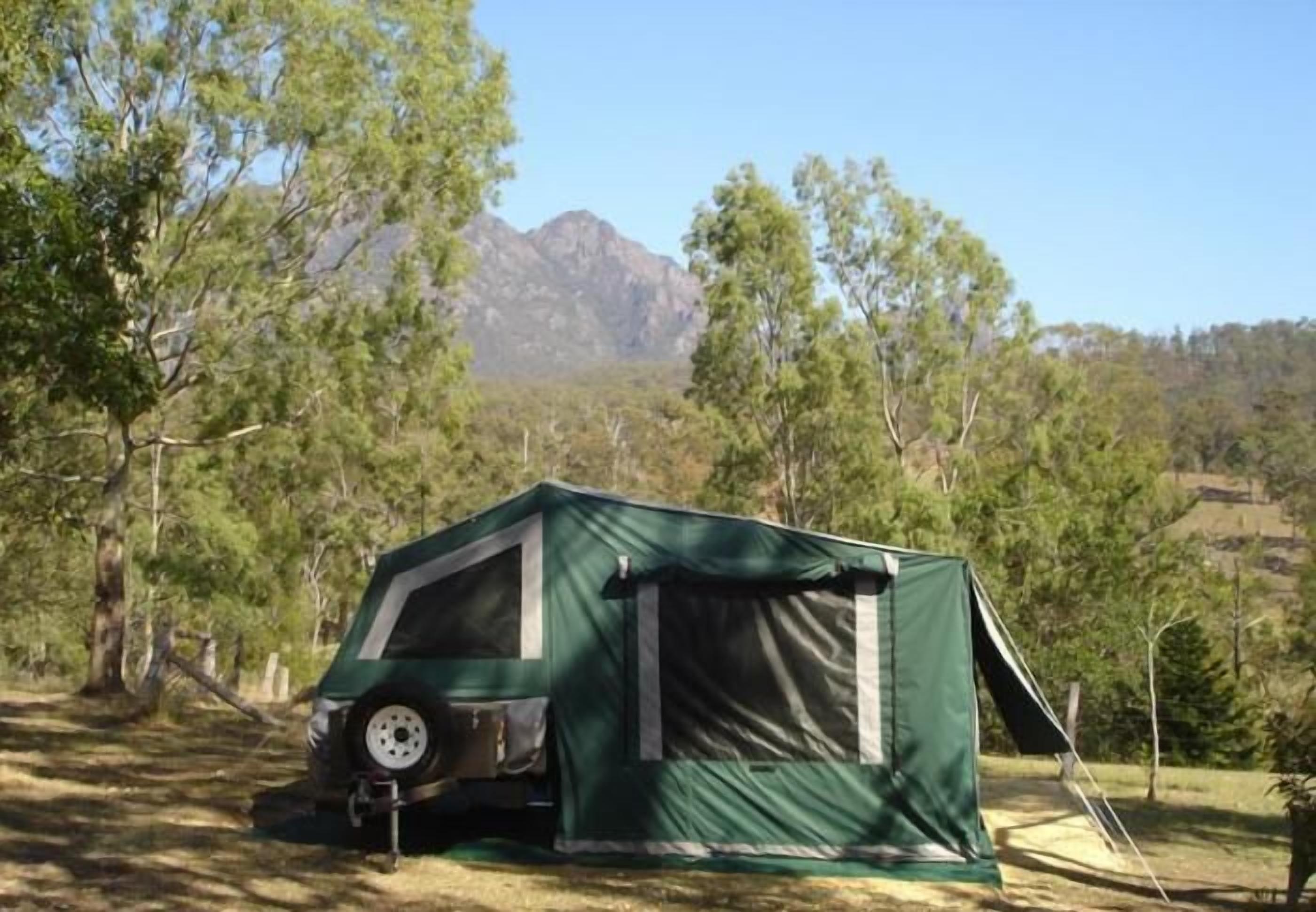 A green tent with a vehicle parked in front, set in a forested area with trees and a mountain in the background.