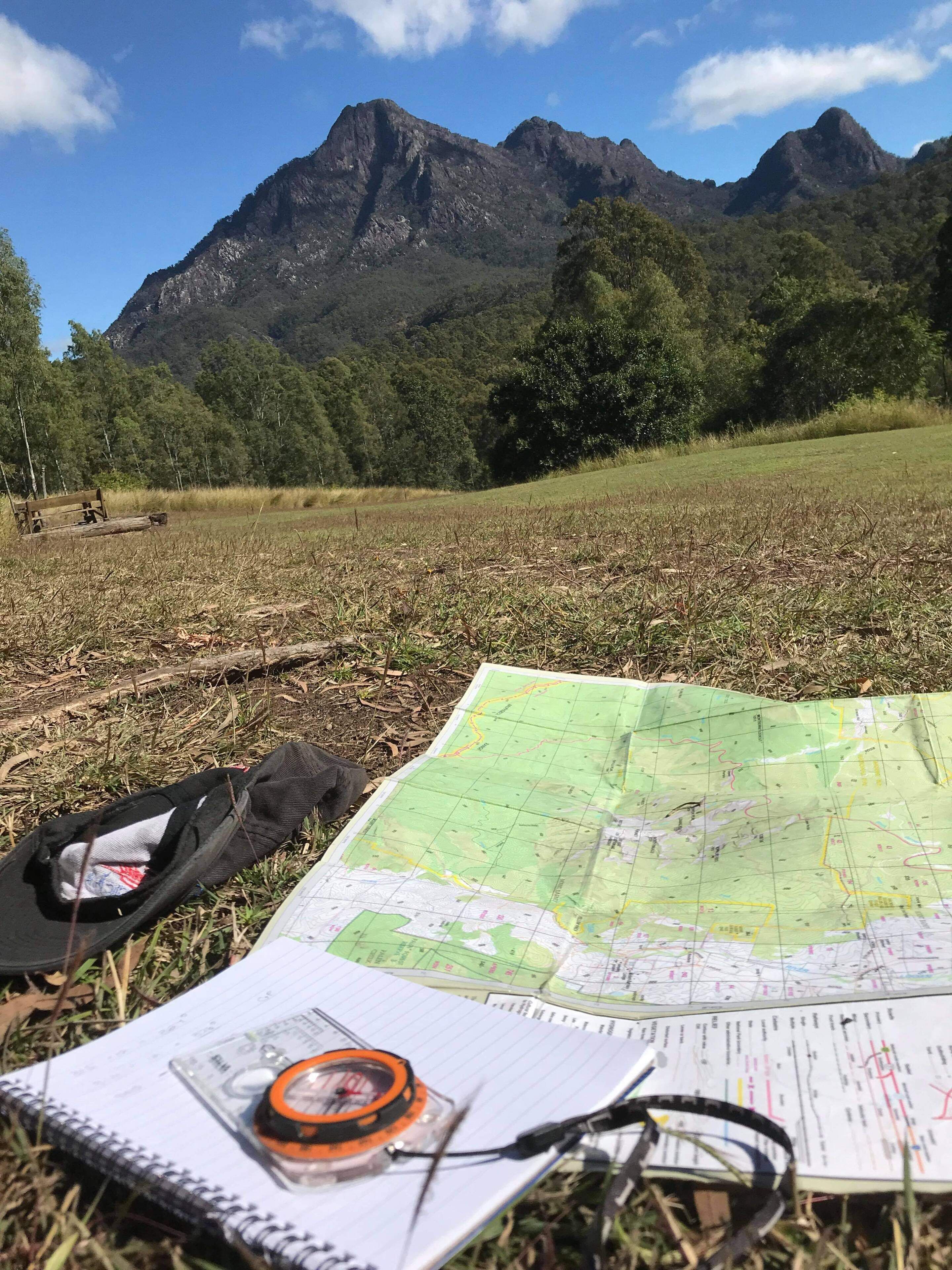 A map, compass, and notebook on grass with mountains in the background.