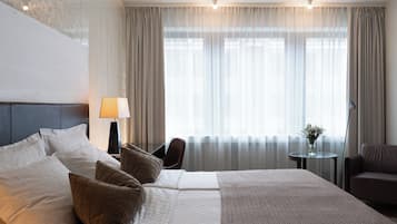 Une chambre d’hôtel avec un grand lit, une table de chevet avec une lampe, une chaise, une petite table avec un vase et une vue sur l’extérieur à travers des rideaux légers.