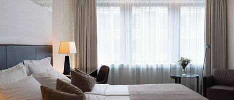 A hotel room with a large bed, a bedside table with a lamp, a chair, a small table with a vase, and a view of the outside through sheer curtains.