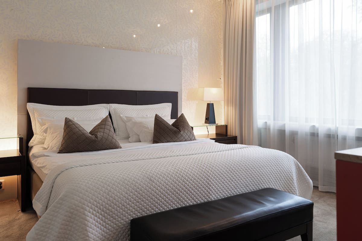 A hotel room with a large bed, a bedside table with a lamp, and a window with sheer curtains.