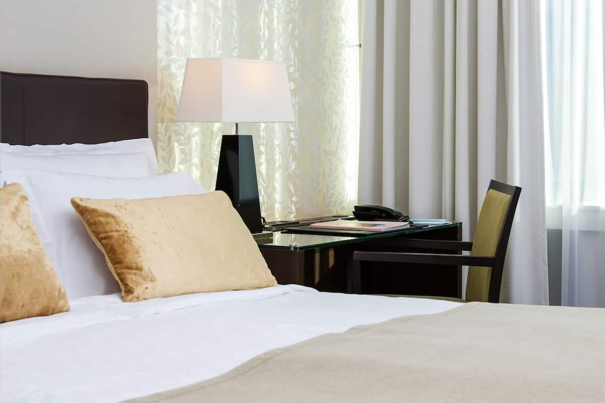 A hotel room with a bed, a desk with a lamp, a chair, and a window with curtains.