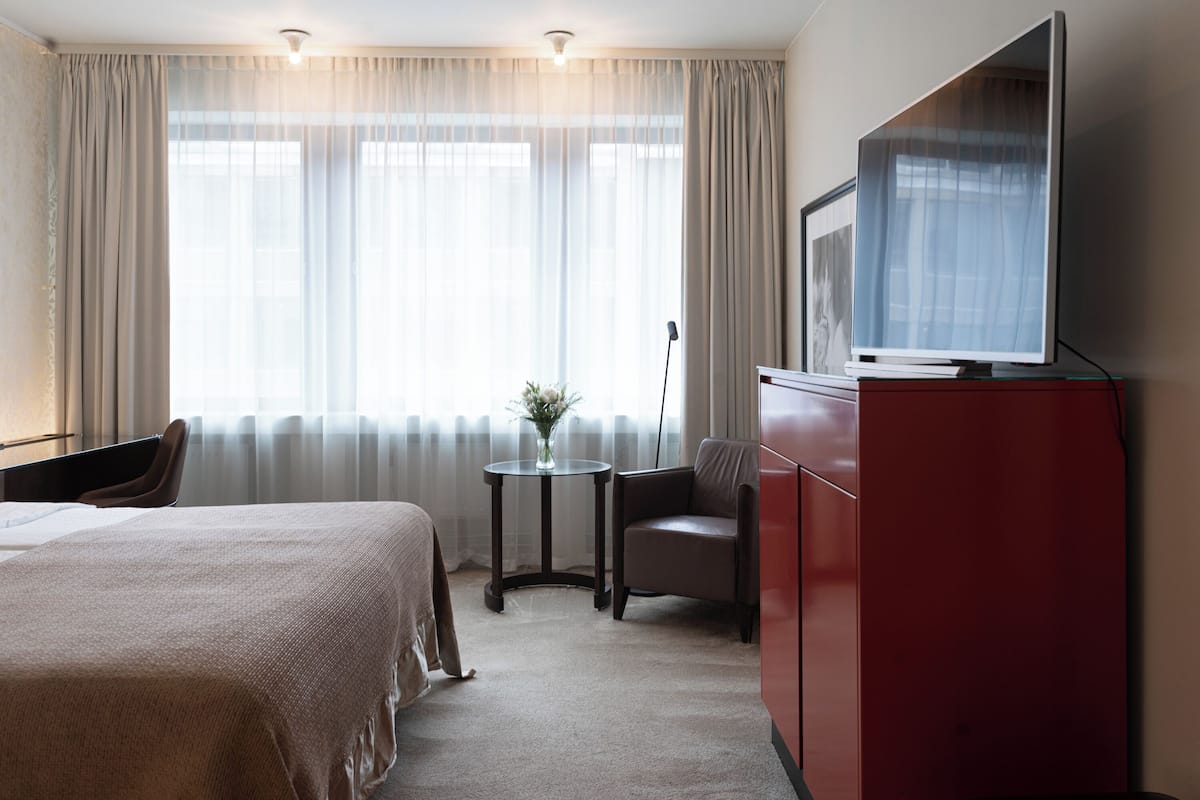 A hotel room with a bed, a TV on a cabinet, a small table with a vase, and a chair.