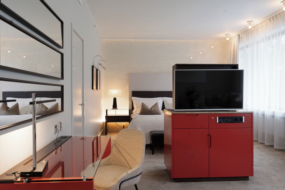 A hotel room with a bed, a TV on a red cabinet, a chair, a desk, and a lamp.
