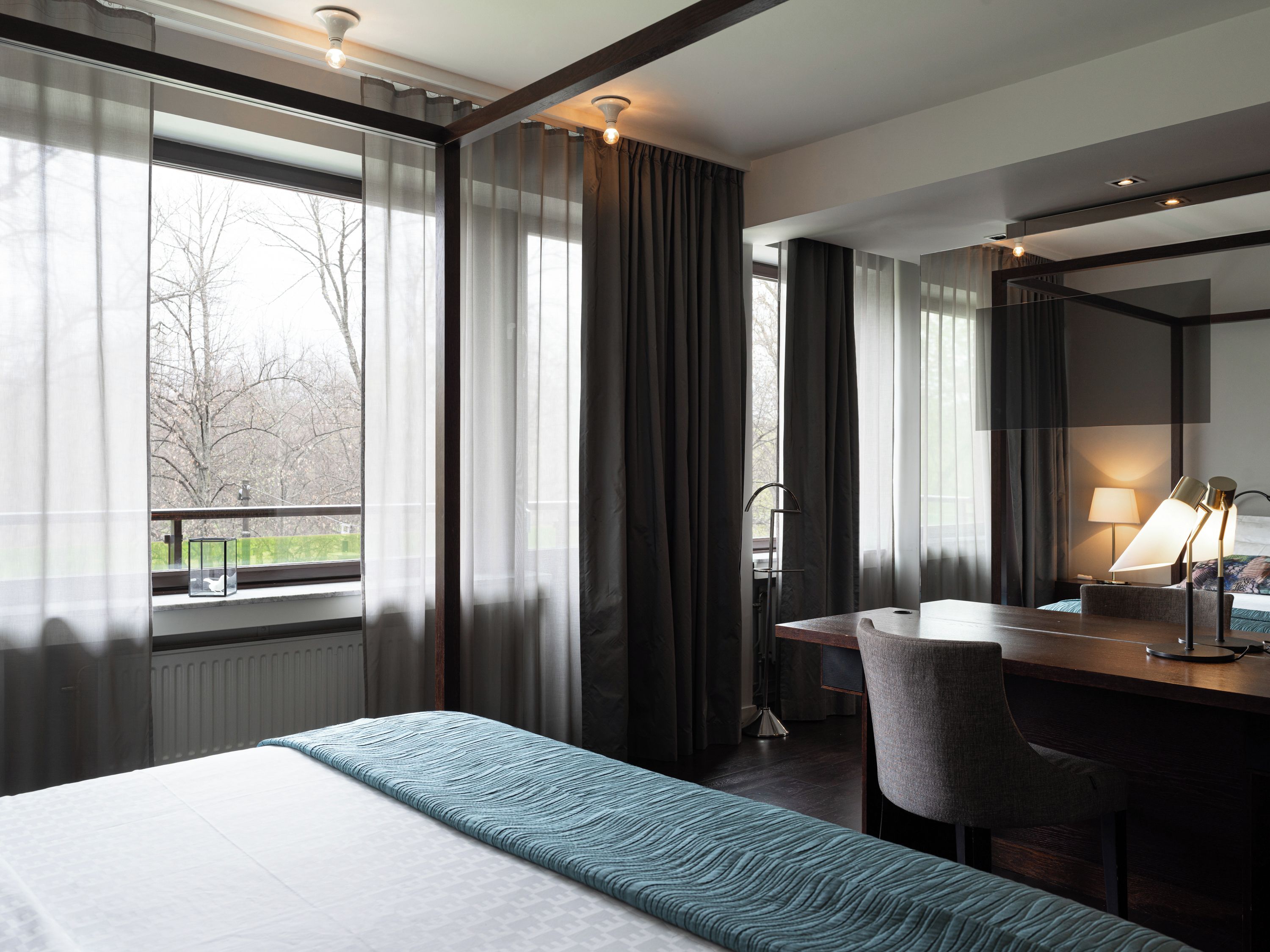 Quarto de hotel com cama, escrivaninha, cadeira, luminária e vista para árvores pela janela.