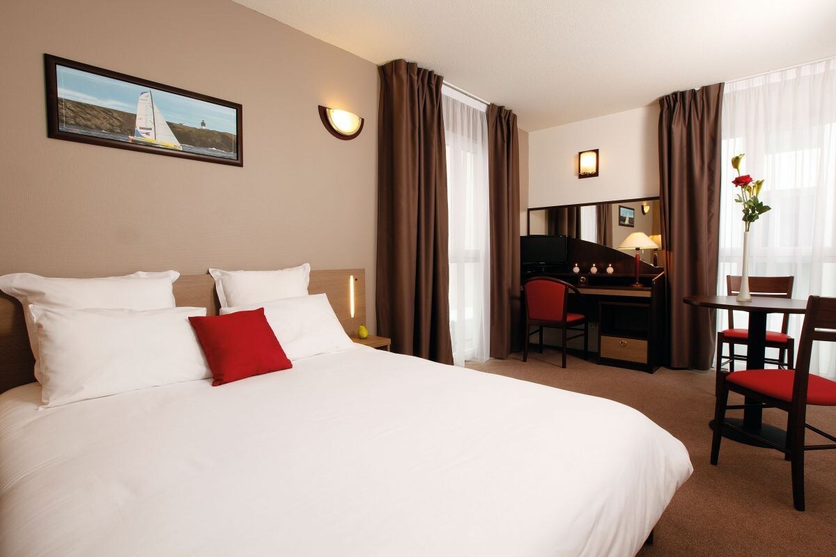 A hotel room with a large bed, a desk with a chair, a TV, and a small table with a vase of flowers.