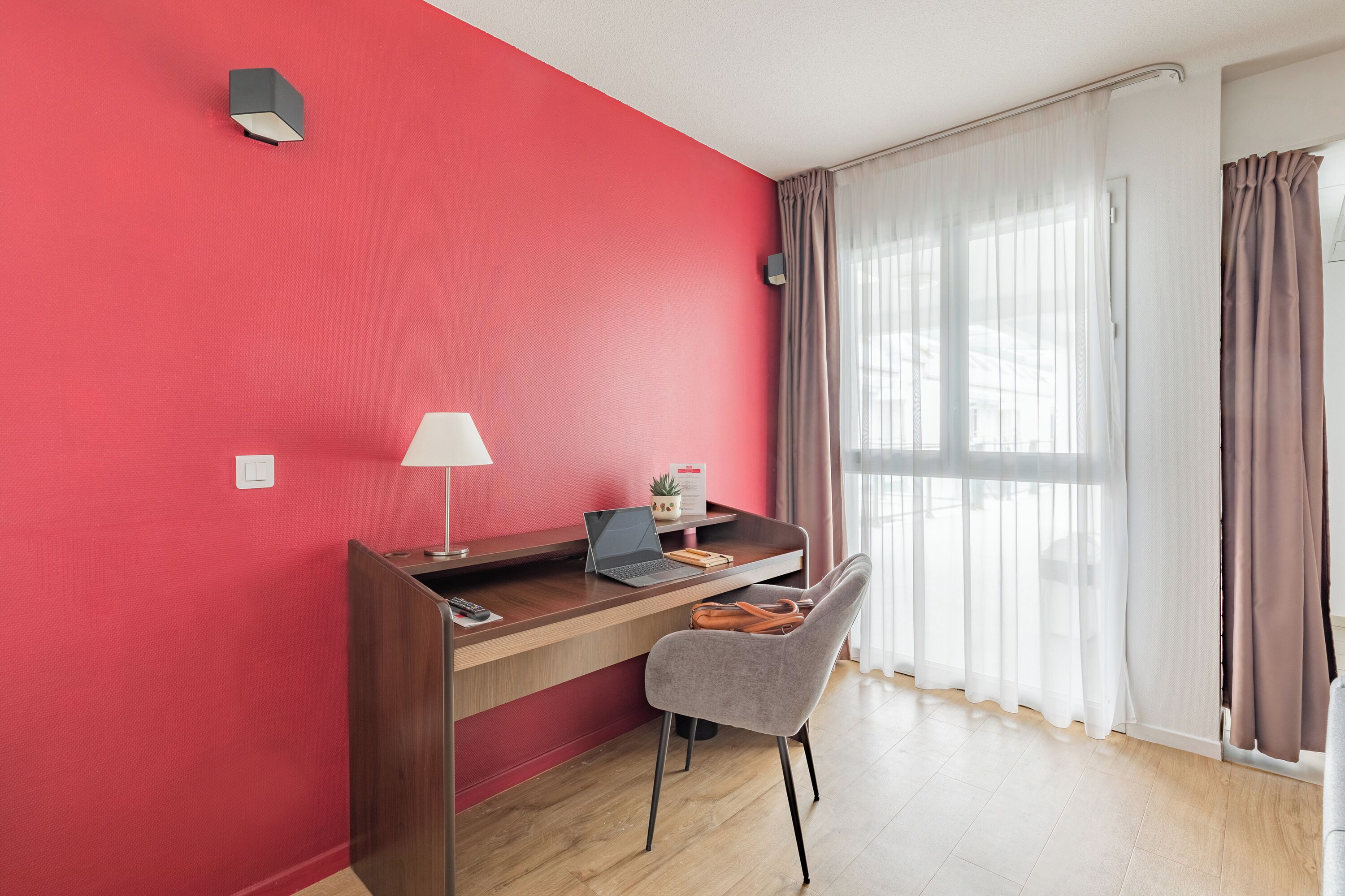 A room with a red wall, a desk with a laptop, a chair, a lamp, and a window with curtains.