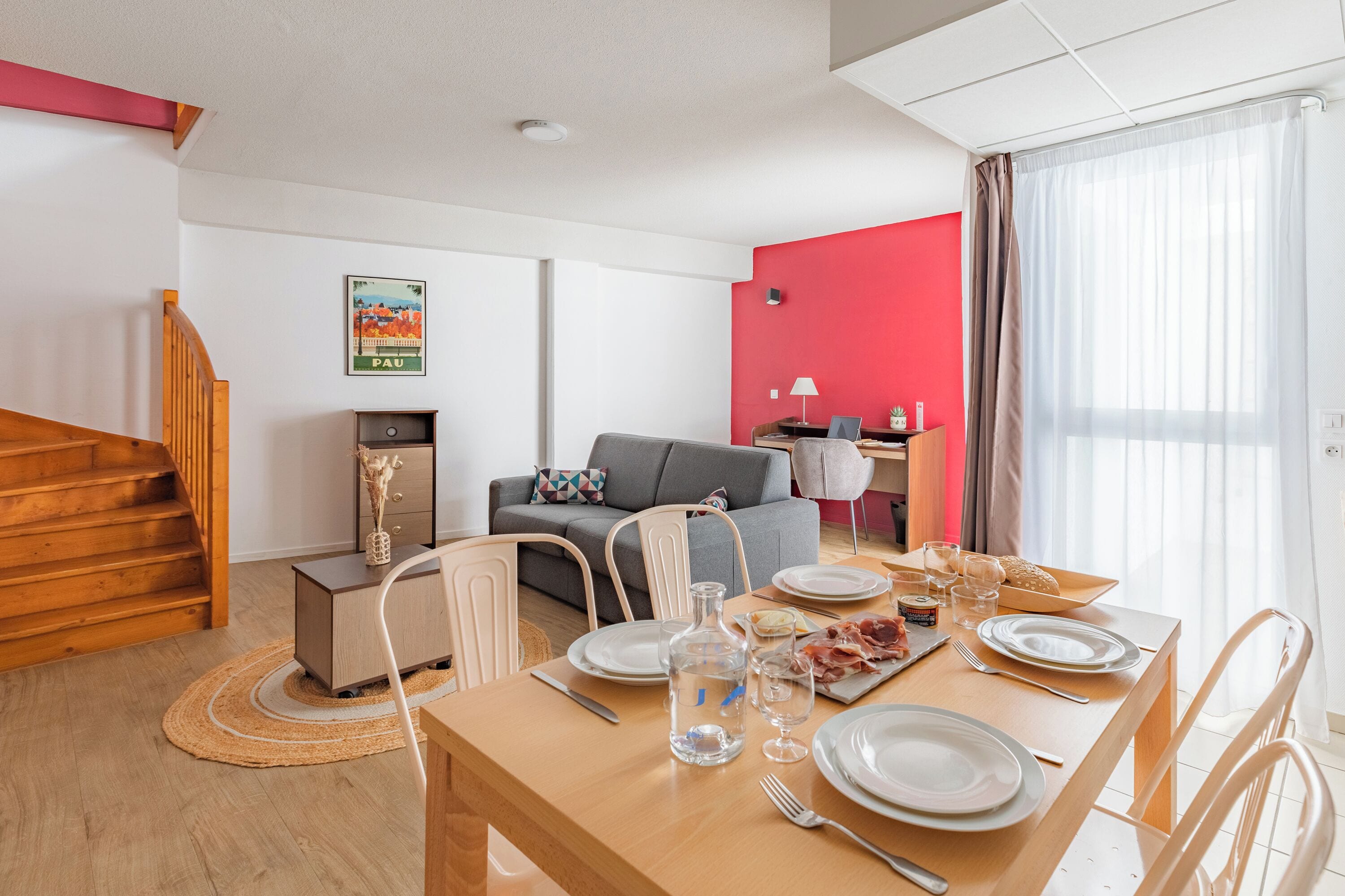 A dining area with a wooden table set for two, a grey sofa, and a staircase with wooden railings.